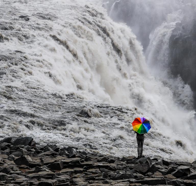 tosender Wasserfall mit Person und Regenschirm mit Regenbogenfarben